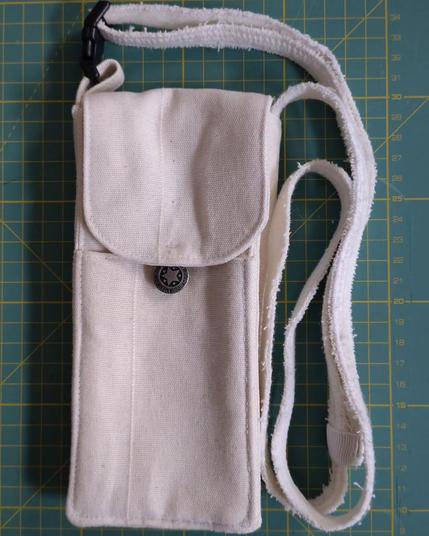 Sacoche pour téléphone mobile faite avec des restes de rideau blanc avec une sangle et rabat avec un bouton en métal (comme ceux des jeans), une ficelle permet de fermer le rabat sur le bouton. Elle est posée sur mon tapis de coupe classique (il est vert et on voit les lignes jaunes qui indiquent chaque centimètre)