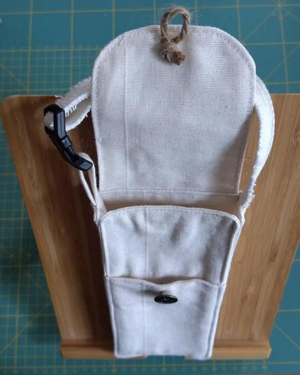 Sacoche pour téléphone mobile faite avec des restes de rideau blanc avec une sangle et rabat avec un bouton en métal (comme ceux des jeans), une ficelle permet de fermer le rabat sur le bouton. Elle est ouverte, on voit qu'elle a une poche extérieure. Elle est posée sur un support en bois de bambou qui est sur mon tapis de coupe classique (il est vert et on voit les lignes jaunes qui indiquent chaque centimètre)