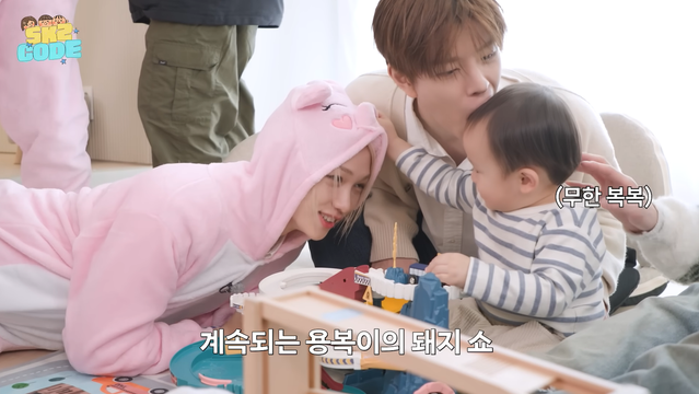 Felix (wearing a pig onesie) smiles at a baby who is reaching out to touch his head. Seungmin sits behind the baby and attempts to chomp the baby's head.