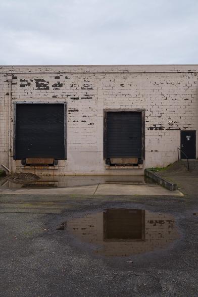 A weathered white brick wall with chipped paint, featuring two closed black roll-up doors and a smaller black entrance door. A large puddle in the foreground reflects the building.