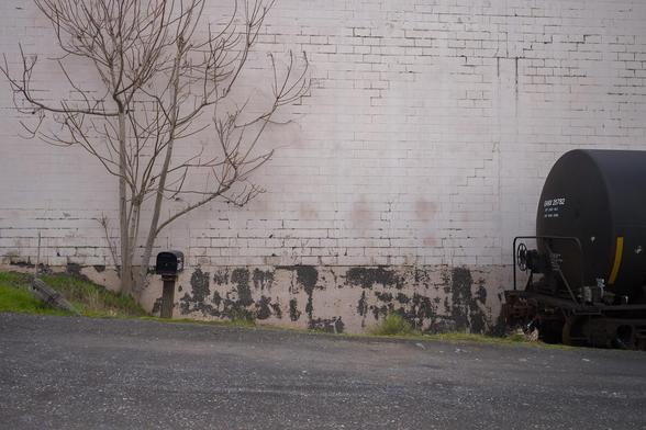 A tree without leaves stands next to a weathered brick wall with peeling paint. There's a mailbox on a post in front of the wall, and part of a black cylindrical railcar is visible on the right side.
