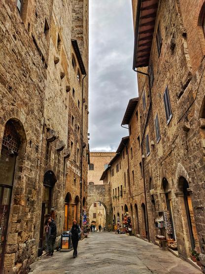 A picturesque, narrow cobblestone street in the historic town of San Gimignano, Italy. The scene is framed by tall, rustic stone buildings with arched doorways and shuttered windows, showcasing the town’s medieval architecture. People stroll leisurely along the street, which gently slopes upward, leading the eye toward an ancient stone archway in the distance. The sky above is overcast, adding a soft, muted light to the warm tones of the buildings. Shops and small businesses line the street, inviting visitors to explore.