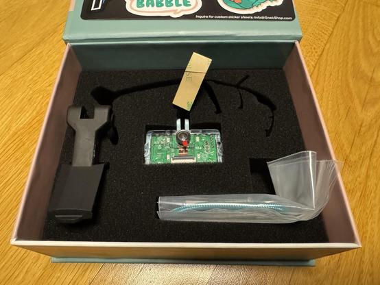 Photo of the inside of the Babble Project box, containing a tracker with a transparent shell with a PCB and a camera on it. A mounting bracket, some cabling, stickers and an antenna.