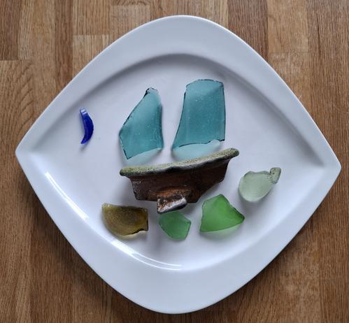 Auf einem weißen Teller liegen verschiedene Scherbenfundstücke.
Ein Teil eines alten Tonkruges, welches mich an den Korpus eines Bootes erinnert.
2 große türkisfarbene Scherben, die als Segel fungieren.
Eine blaue Mondsichel.
Unter dem "Boot" veir Scherben in unterschiedlichen Grüntönen, die an Wellen erinnern.