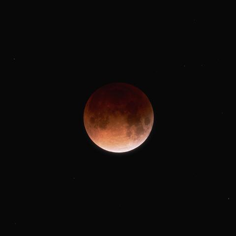 A reddish, partially shadowed moon during a lunar eclipse hangs in a black sky with a few faint stars visible around it.