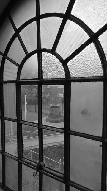 Black and white photo, a large window fills the frame, viewed from inside the building. Made of many panes separated by ironwork, the window is semicircular at the top, the iron work radiating out like sun rays. A square of four panes in the middle is in clear glass, with a handle suggesting that this section is openable. The other panes are a variety of different textured frosted glass. Through the clear panes the courtyard outside can be seen, and a large bell cope, used for casting bells, is in the courtyard.