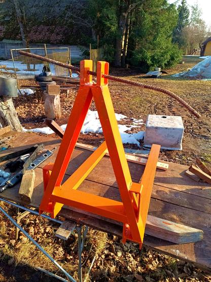 Self-made pallet forks for a mini-tractor's rear attachment sitting on two wooden spacers on top of an outdoor workbench. They're now painted in bright orange paint, some primer still shining through this first coat. A rusty piece of bent rebar is stuck through the top, the other fittings are removed for painting.