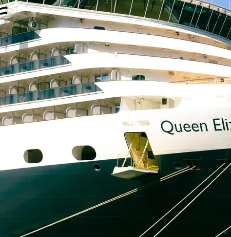 Close-up of a cruise ship’s bow showing stacked balcony decks above a black hull, an open side door with a small platform, thick mooring lines stretching diagonally, and the partial name “Queen Eli” painted on the white side.
