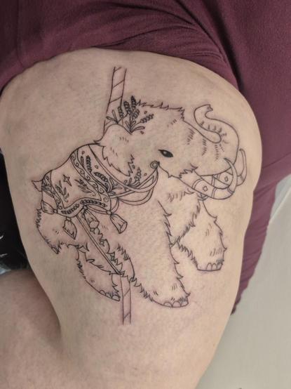 Photo of a freshly done work in progress tattoo of a wooly mammoth done up to look like a carousel ride. It has a blanket type saddle, with embellishments and tassles, with sprigs of lavender as painted designs on it, as well as some sprigs tucked behind its ear. It is very wooly and fluffy and looks happy doing a big steppy.