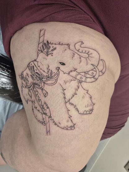 Photo of a freshly done work in progress tattoo of a wooly mammoth done up to look like a carousel ride. It has a blanket type saddle, with embellishments and tassles, with sprigs of lavender as painted designs on it, as well as some sprigs tucked behind its ear. It is very wooly and fluffy and looks happy doing a big steppy. This time at a different angle.