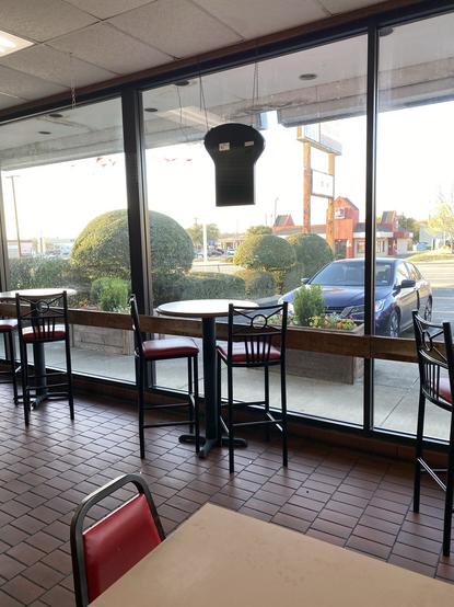 The image features the interior of a fast-food restaurant with a view of the outside. There's a table and several high-backed chairs, mostly in red and black. Large windows provide natural light and showcase landscaped bushes and a parking lot beyond. A hanging sign blocks the view a bit. Jack in the Box across street