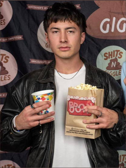 Young Hudson Williams with a blank expression wearing a leather coat and white t shirt. In front of a banner with logos that say "shits n giggles". Holding a soda and a bag of popcorn but the popcorn font really looks like it says poop. Middle fingers slightly out