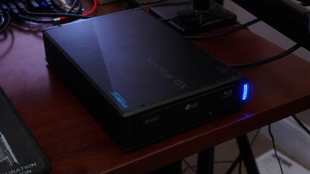 An LG WH16NS60 optical drive in an NexStar DX 5.25" USB3 enclosure sitting on top of a desk. A violently bright blue LED flashes showing disc reading activity.