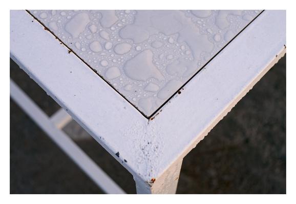 The corner of a white table covered in water droplets, showing slight wear and rust on the painted metal frame. - Google Gemini 3.1 Pro Preview