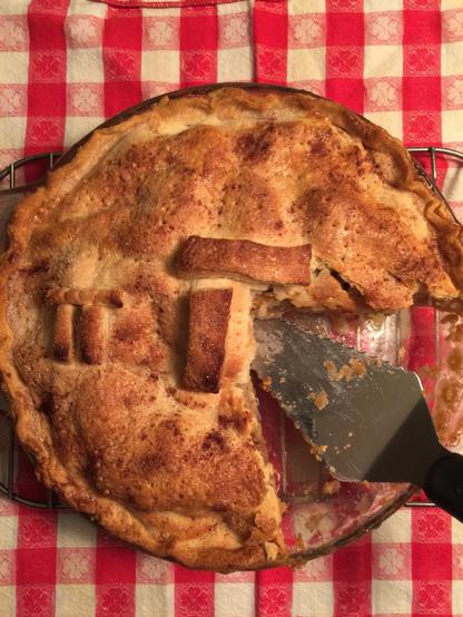 Color image of today's Pi Pie for Pi Day. Image shows a home made apple pie with a Pi symbol on top, on a red gingham tablecloth, a pie slicer having lifted out a piece.