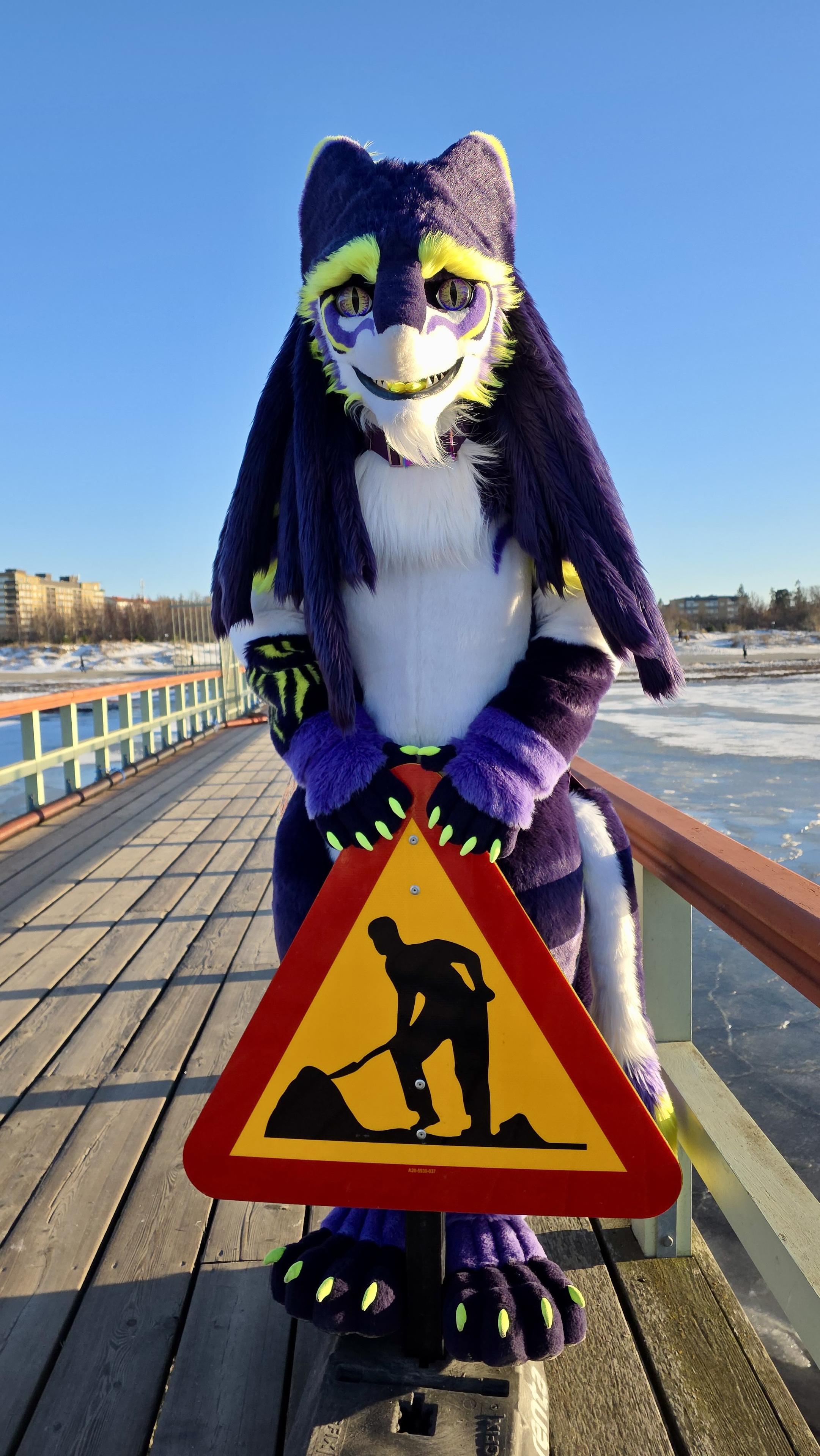 A Sergal stands on a wooden pier, holding a construction warning sign. The background features a clear blue sky and buildings, with some ice visible on the water.