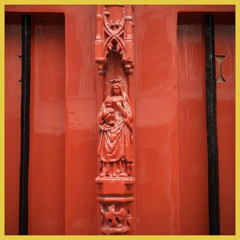 Detail of a church door in old red, with a fully integrated statue of Mary with child in the middle of the double door. To the left and right of it 2 black bars. The whole scene is evenly lit.