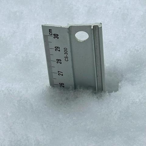 ein Messstab steckt senkrecht im frisch gefallenen Schnee. Der Schnee reicht auf der Skala bis an die 26 cm.