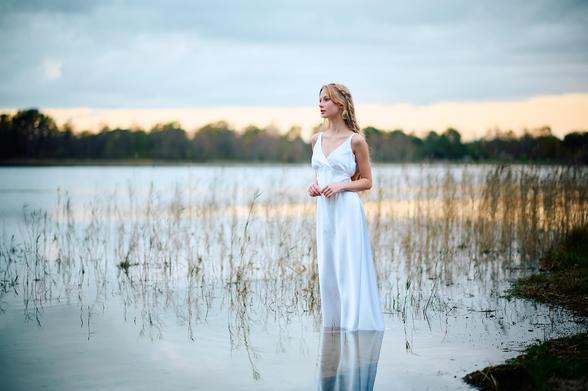 Young scandinavian model with long, blonde hair, standing in the shallow waters near the brink of a still lake in the sunset in a white, flowy dress.