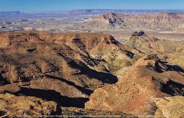 View overlooking a canyon network of multicolored soils and rocks in a desert setting; a narrow dirt road can just be seen winding down the canyon. Beyond a basin lie other finger mesas and in the distance a sandstone cliff. Hazy blue sky.
©BosqueBill.com