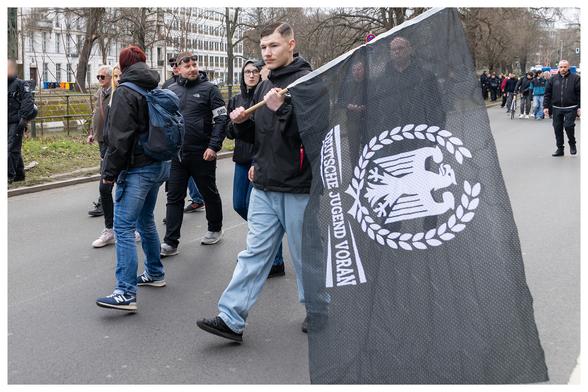 Querformat Bild mit einem kleinem weißen Rahmen um das Foto.
Ein junger Neonazi mit Seitenscheitel, schwarzer Jacke und Blue Jeans trägt eine 'Deutsche Jugend voran' Fahne mit dem Logo der neonazistischen Gruppierung.
Im Hintergrund weitere Neonazis