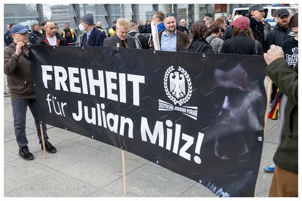 Querformat Bild mit einem kleinen Rahmen um das Foto.
Neonazis hinter einem schwarzen Transparent mit der Aufschrift:"Freiheit für Julian Milz!" mit dem Konterfei von Milz und dem Logo der neonazistischen Gruppierung 'Deutsche Jugendvoran'