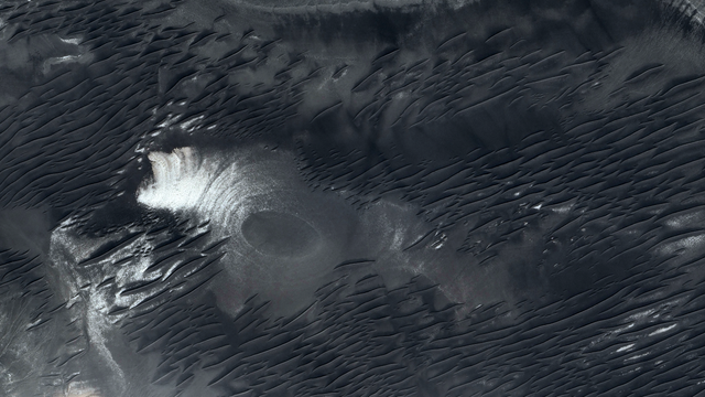 A satellite picture of a desertic landscape with black sand, small dunes and an ovoid shape in the middle.