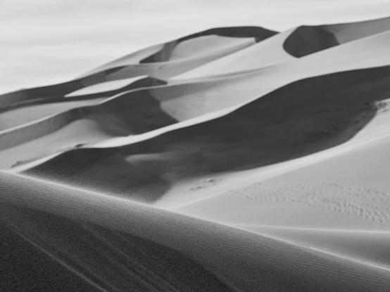 A black and white photograph of sand dunes with shadows and ripples, the sand blowing past in the foreground.