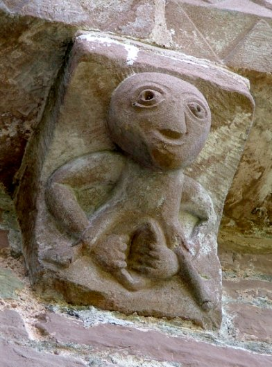A stone carving in a church depicting a bald figure who kind of resembles ET with a large, exaggerated vulva. The figure is holding the vulva open with their hands.