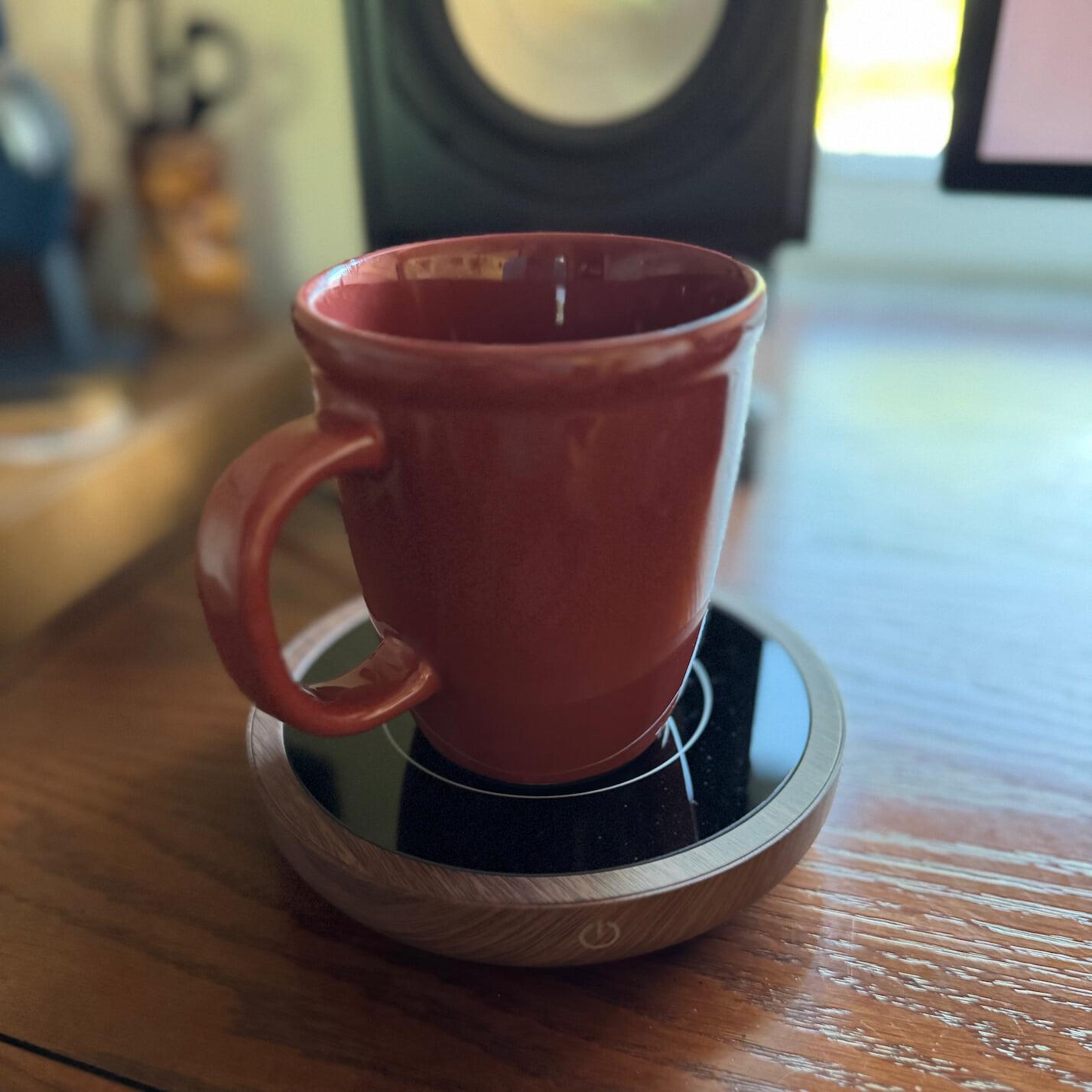 A reddish-brown coffee mug sitting on a small, round mug warming plate, which has a black top and wooden sides. It’s sitting on a wooden desk in front of a speaker.