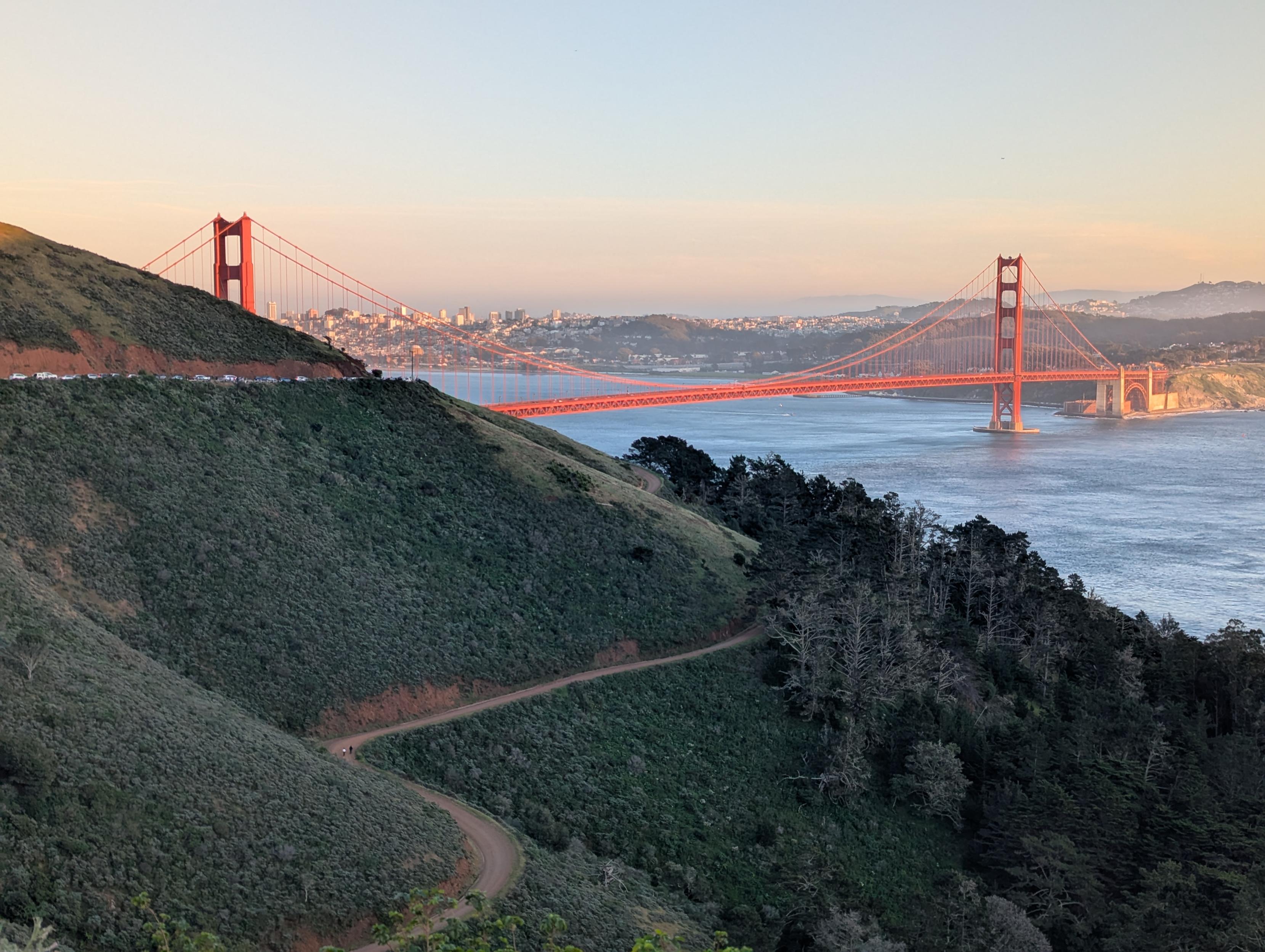 Same photo as before description below but everything turned up to 11 for HDR, color vibrancy, and sharpness.  Photo of the Golden Gate bridge from the north side looking towards San Francisco at sunset. The red color of the bridge is glowing in the golden hour contrasted in the foreground with green slopes of the hillside under shade carved by red unpaved paths. The left tower of the bridge is only partially visible with the city in the distance poking through the gaps in the cables and the valley beyond the city nestled between the span of two towers bridging across the calm waters of the bay reflecting the steely blue of the sky.