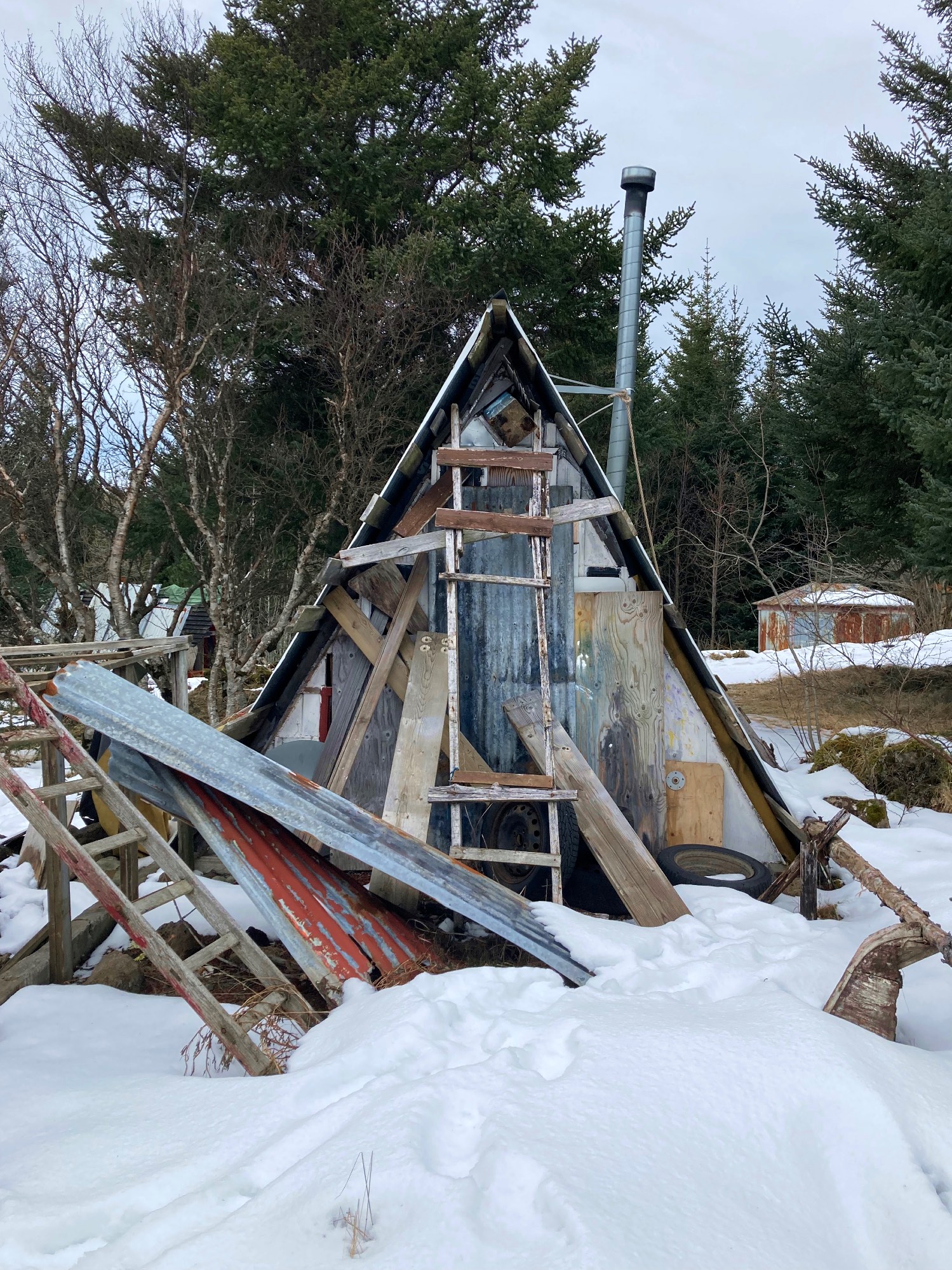An A-frame shed surrounded by trash, snow and trees.