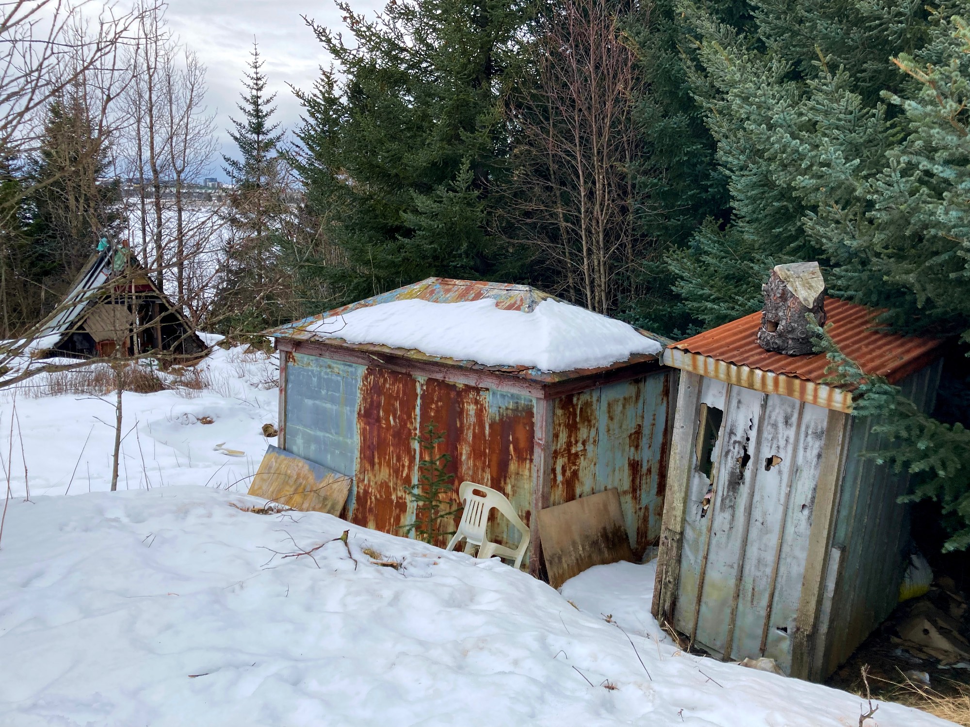 A rusty old shed, an outhouse and an A-frame shed at a distance. There's also a lot of snow and some trees.