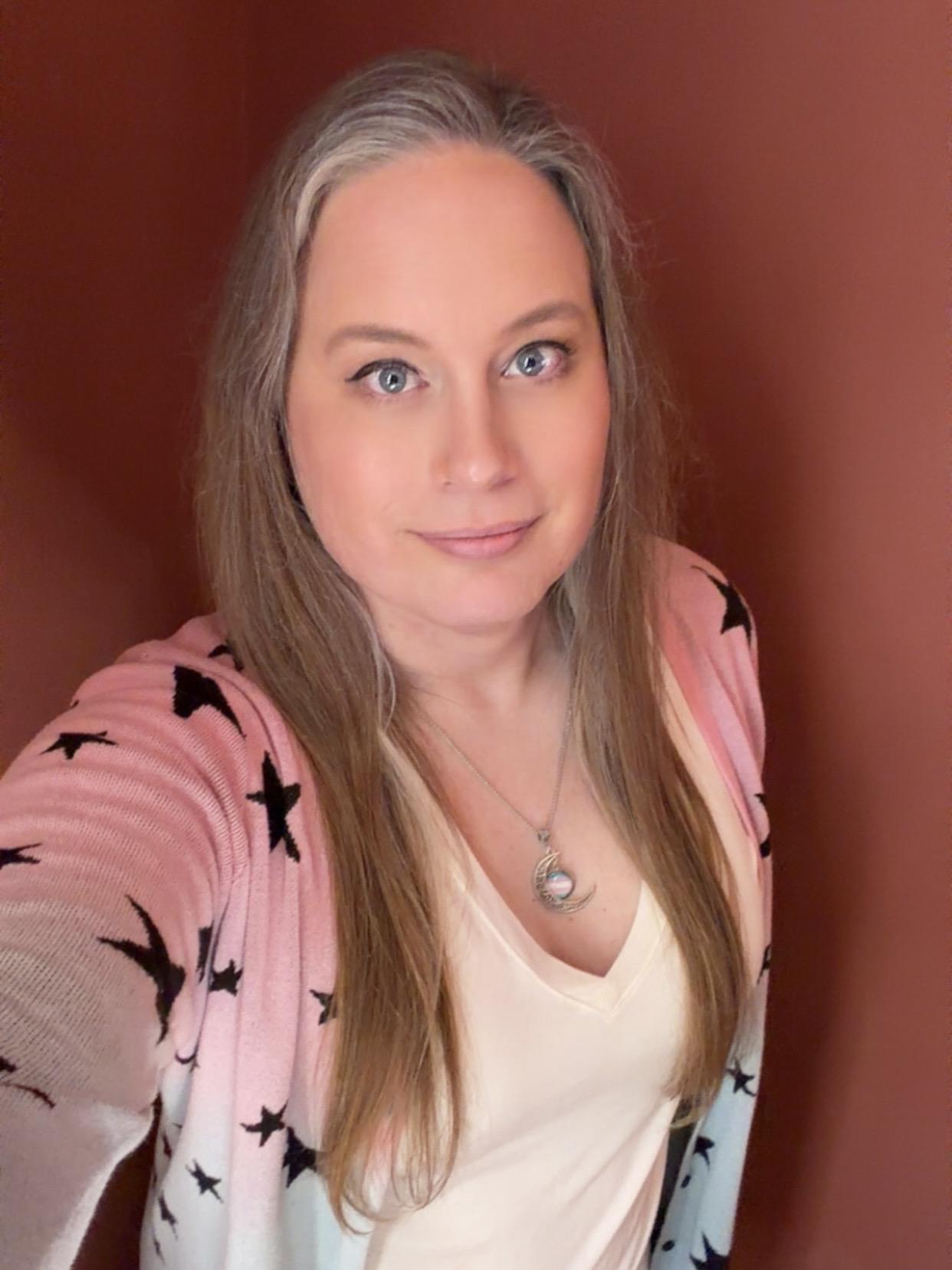 Selfie of a white trans woman wearing pink, white, and blue colors and a trans necklace. Photo was taken with Snapchat to hide wrinkles and electrolysis swelling.