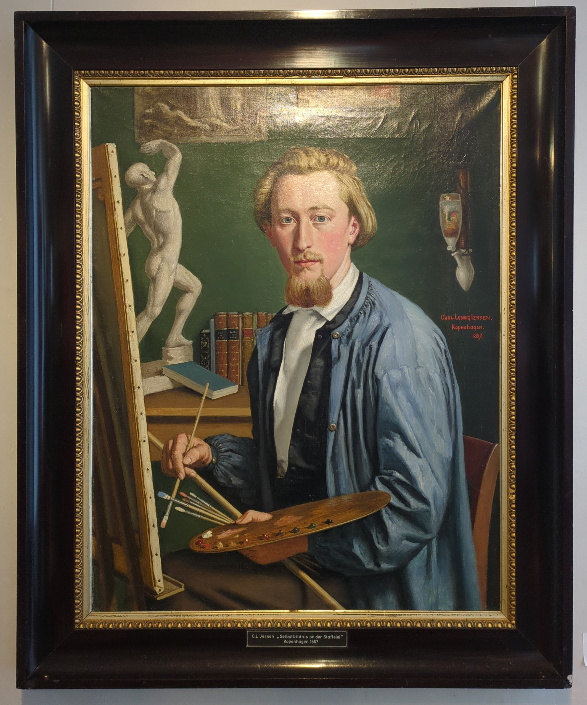 A photo of a painting in a museum in a Husum. The painter painted a selfie of him while painting. He is holding brushes and the palette, the canvas can be seen from the side. The painter himself is looking at you or "in the camera" you could say. The painter is called Carl Ludwig Jessen (1833-1917).