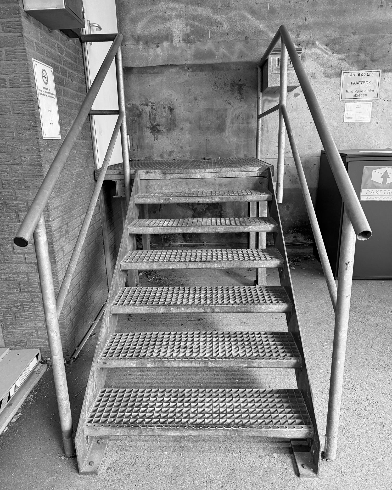 Metalltreppe mit Gitterstufen und beidseitigen Handläufen vor einer Betonwand. Links ist ein Wandkasten und ein Schild zu erkennen, rechts steht ein großer Behälter mit einem Pfeil-Symbol. Der Boden besteht aus glattem Beton.
