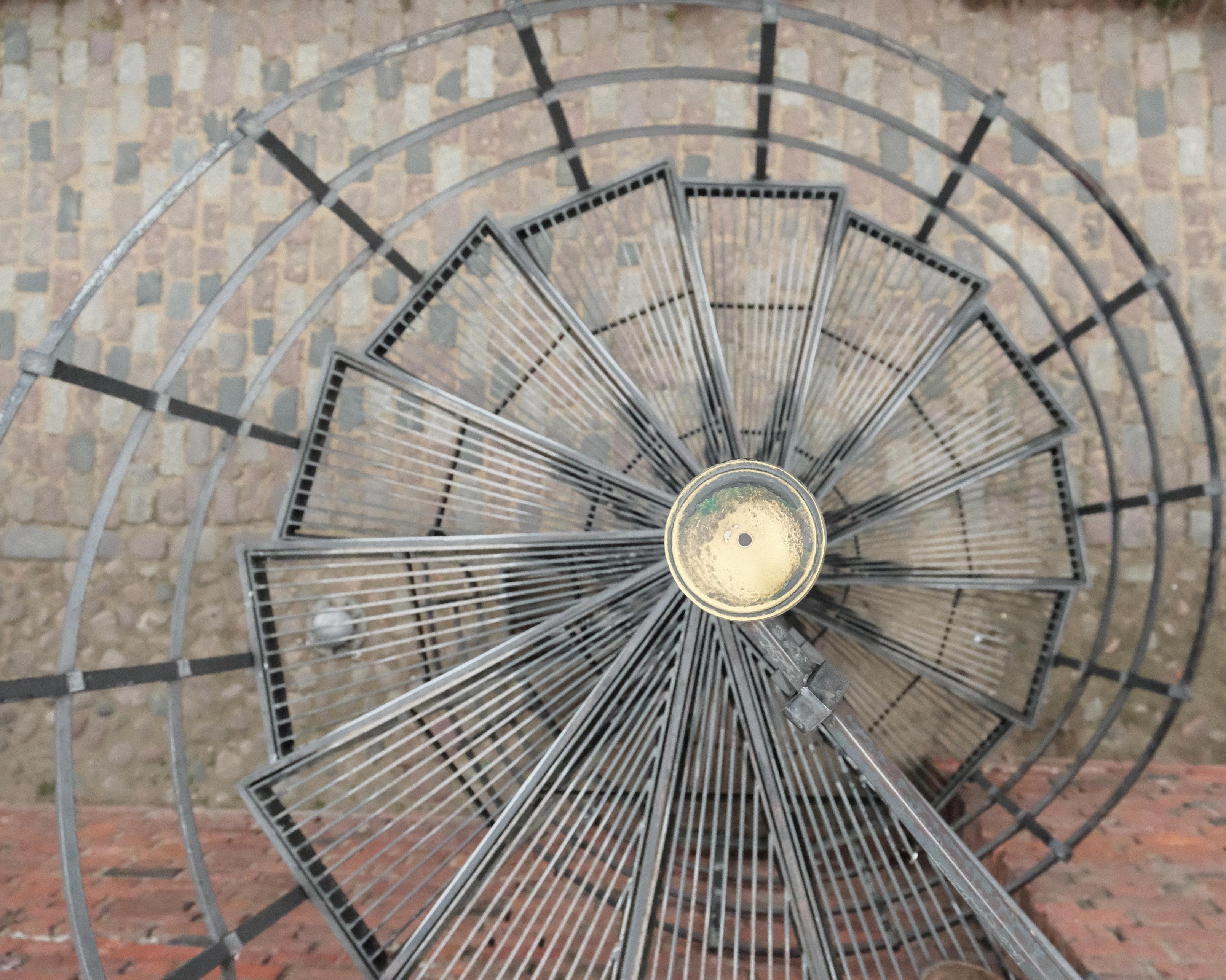 Spiral staircase photographed straight from above in a way that the central column is a focal point, around which the stair is winding. In the background is the stone pavement of the floor, in the lower part of the background there are red bricks of the wall against which the spiral staircase has been built.