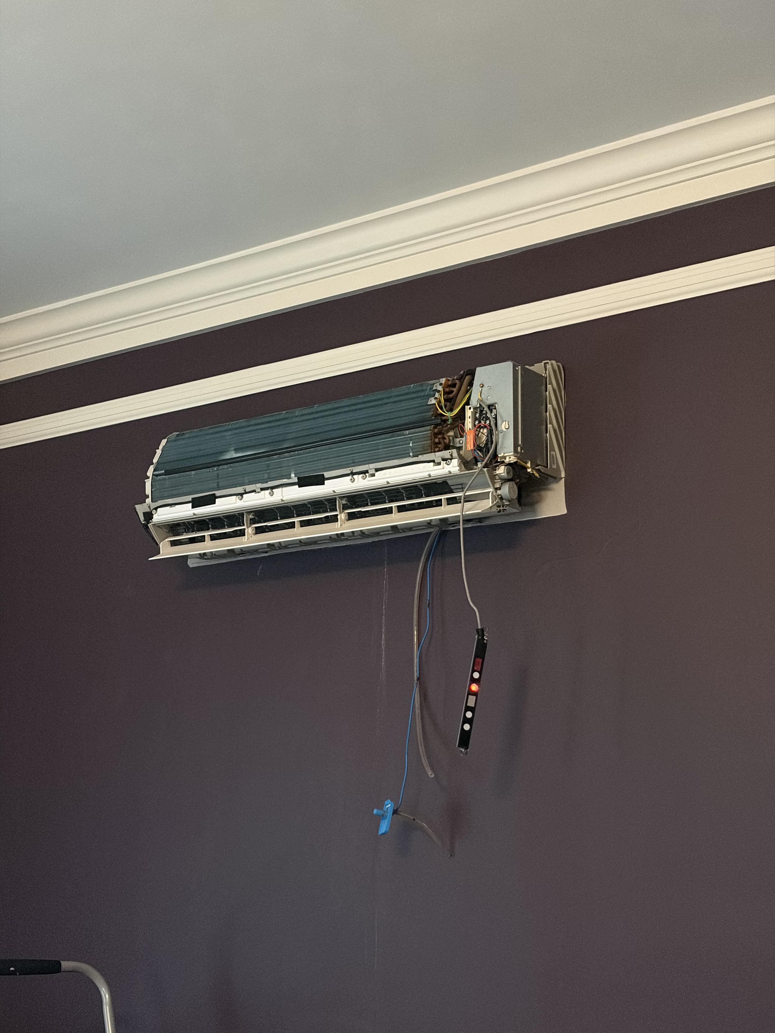 A minisplit air conditioner head unit handing on the wall with the plastic façade off and wires and hoses dangling from the bottom right.