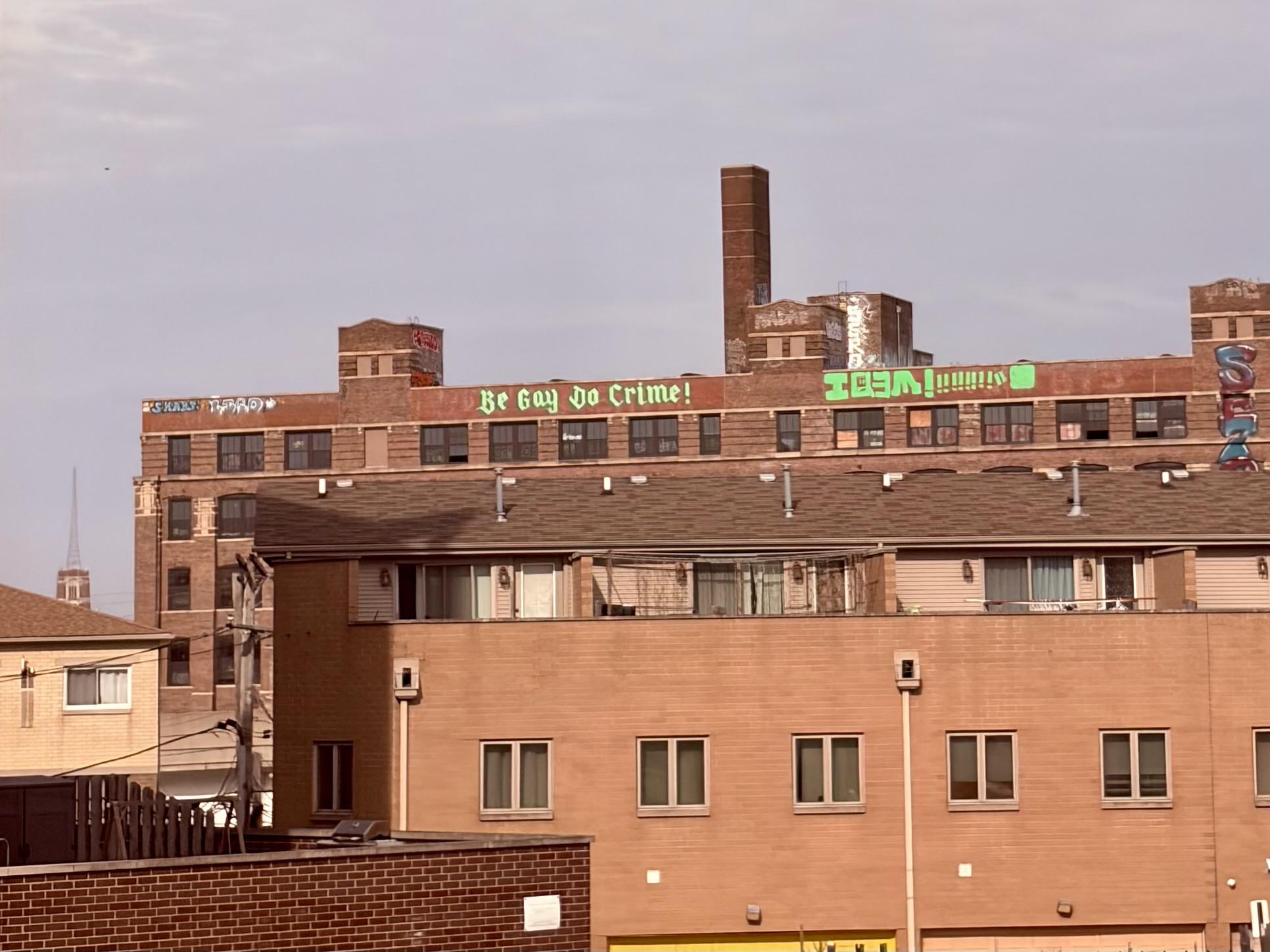 An industrial area of Chicago with “Be Gay Do Crime” spray painted bright green in gothic lettering on the side of brick building large enough to be read several blocks away.