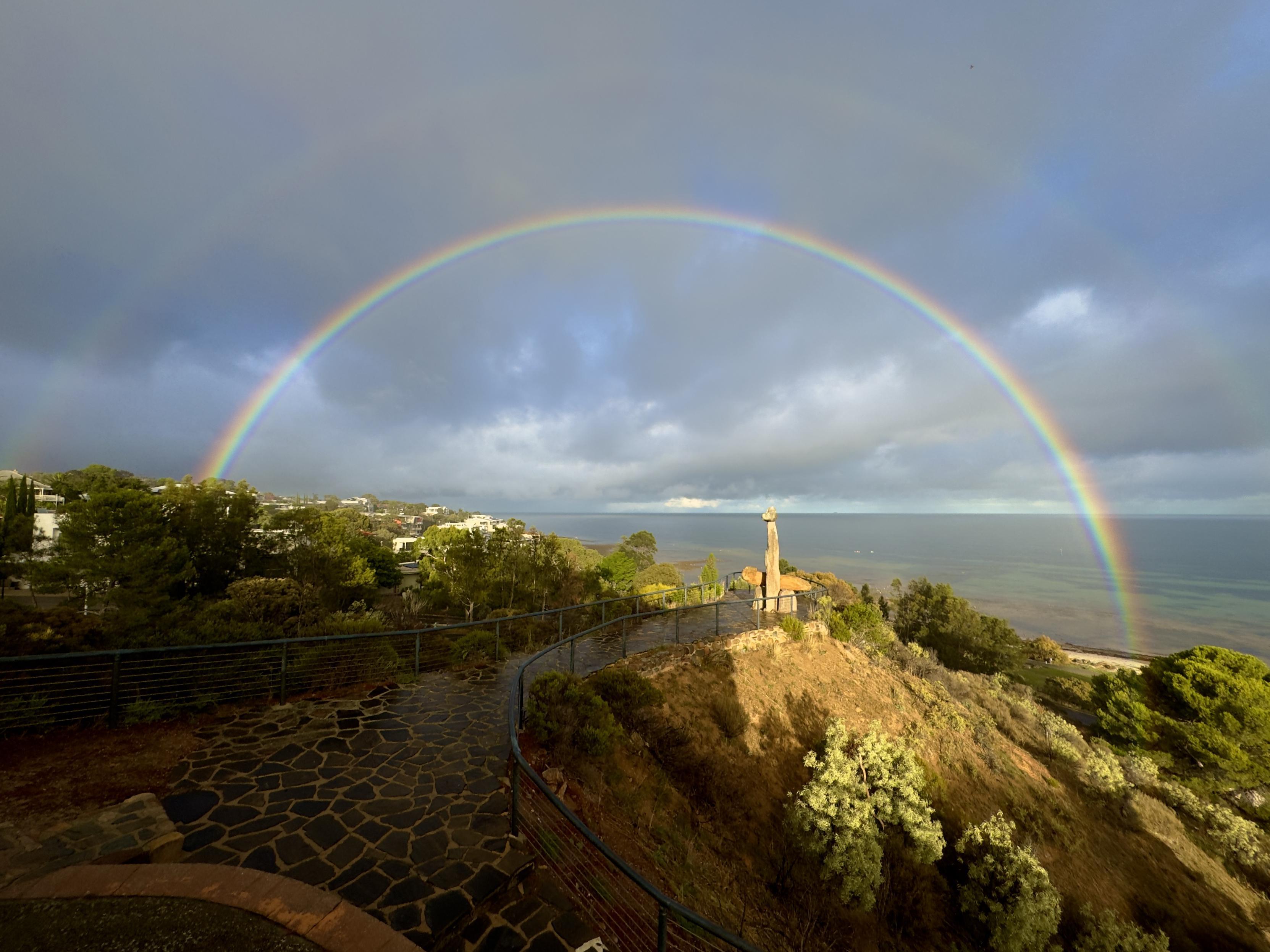 An intense double rainbow over the Tjilbruke Monument in Kingston Park, South Australia. One side of the monument is brightly lit with golden early morning light. The sea looks green and clear today and the pot of gold must be at the popular beachfront café