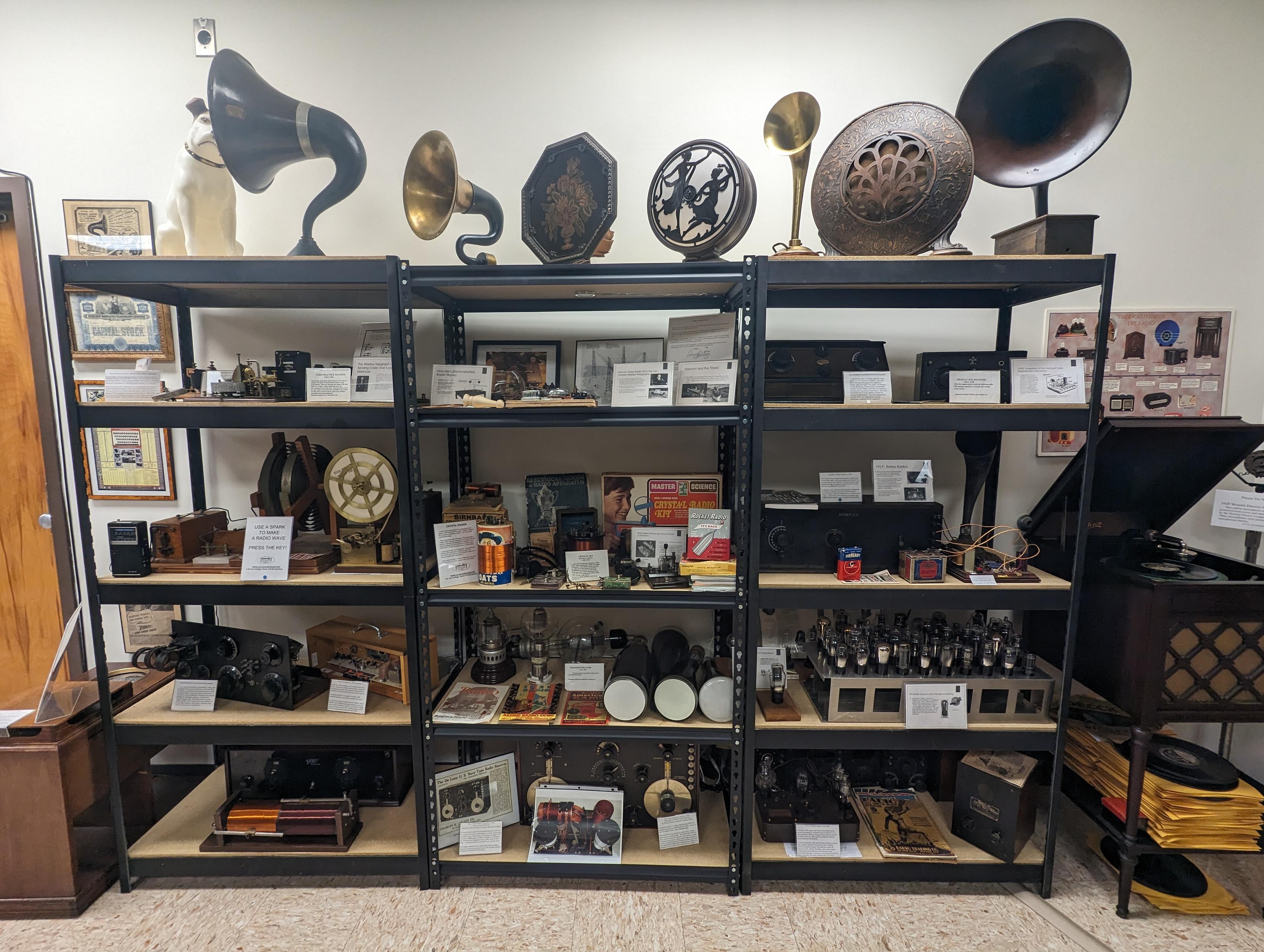 Shelves of older equipment from the beginning of the radio era. Across the top shelves are gramophone horns.