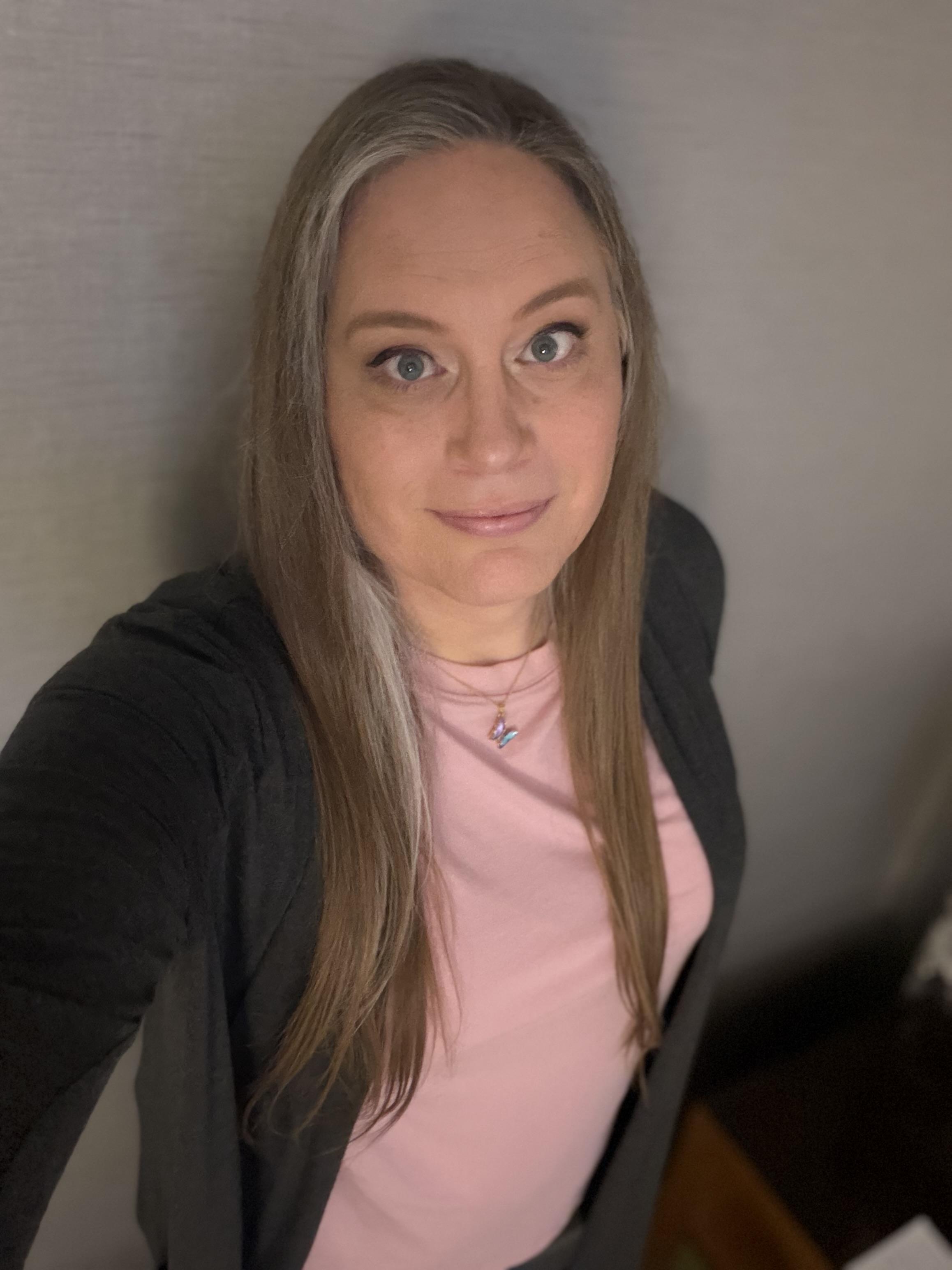 Selfie of a white woman with long hair standing against a wall. She is wearing a pink t-shirt with a gray cardigan top and a butterfly necklace.