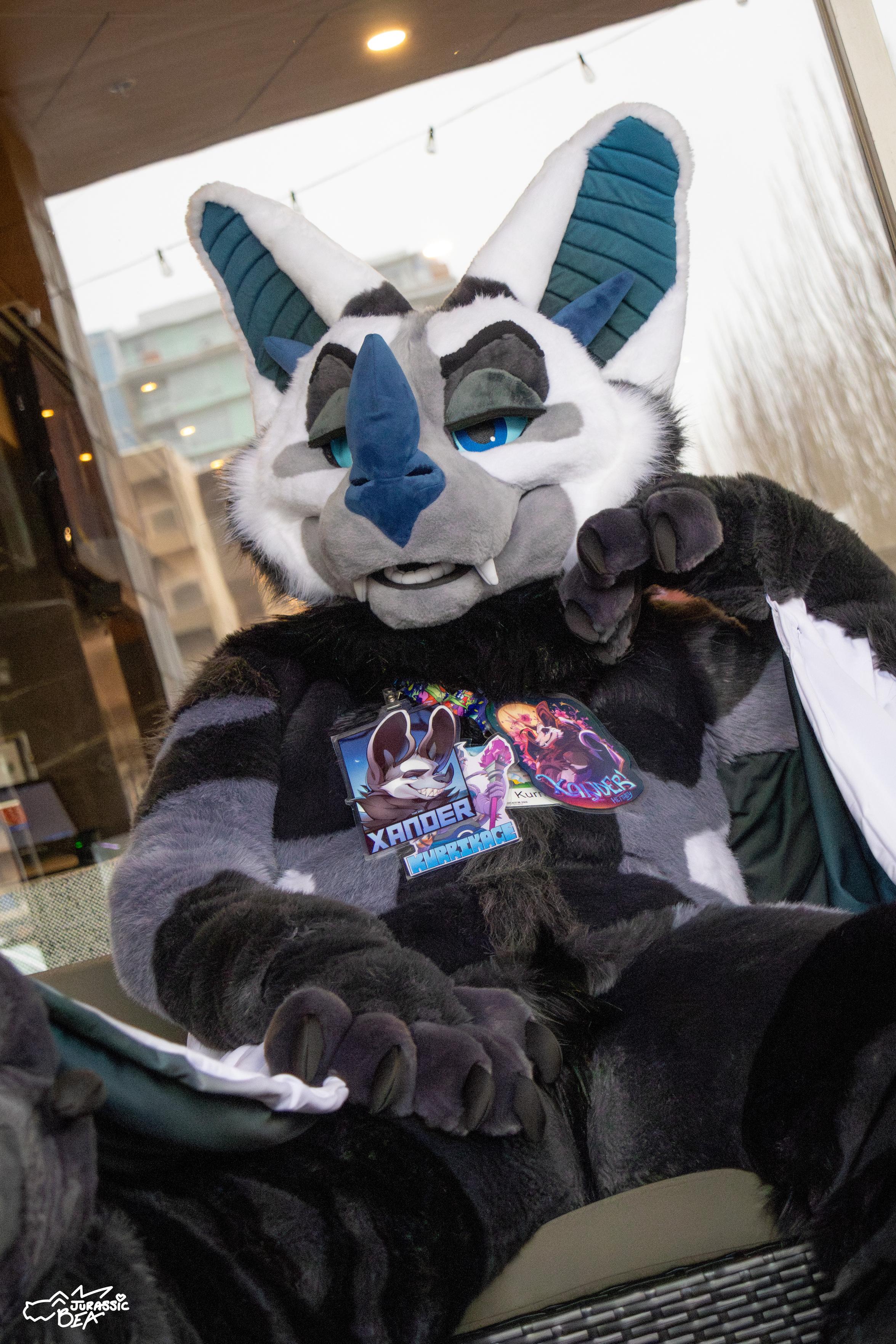 A photo of a bat fursuit sitting on a couch with a leg up and legs spread. Giving the viewer some bedroom eyes.