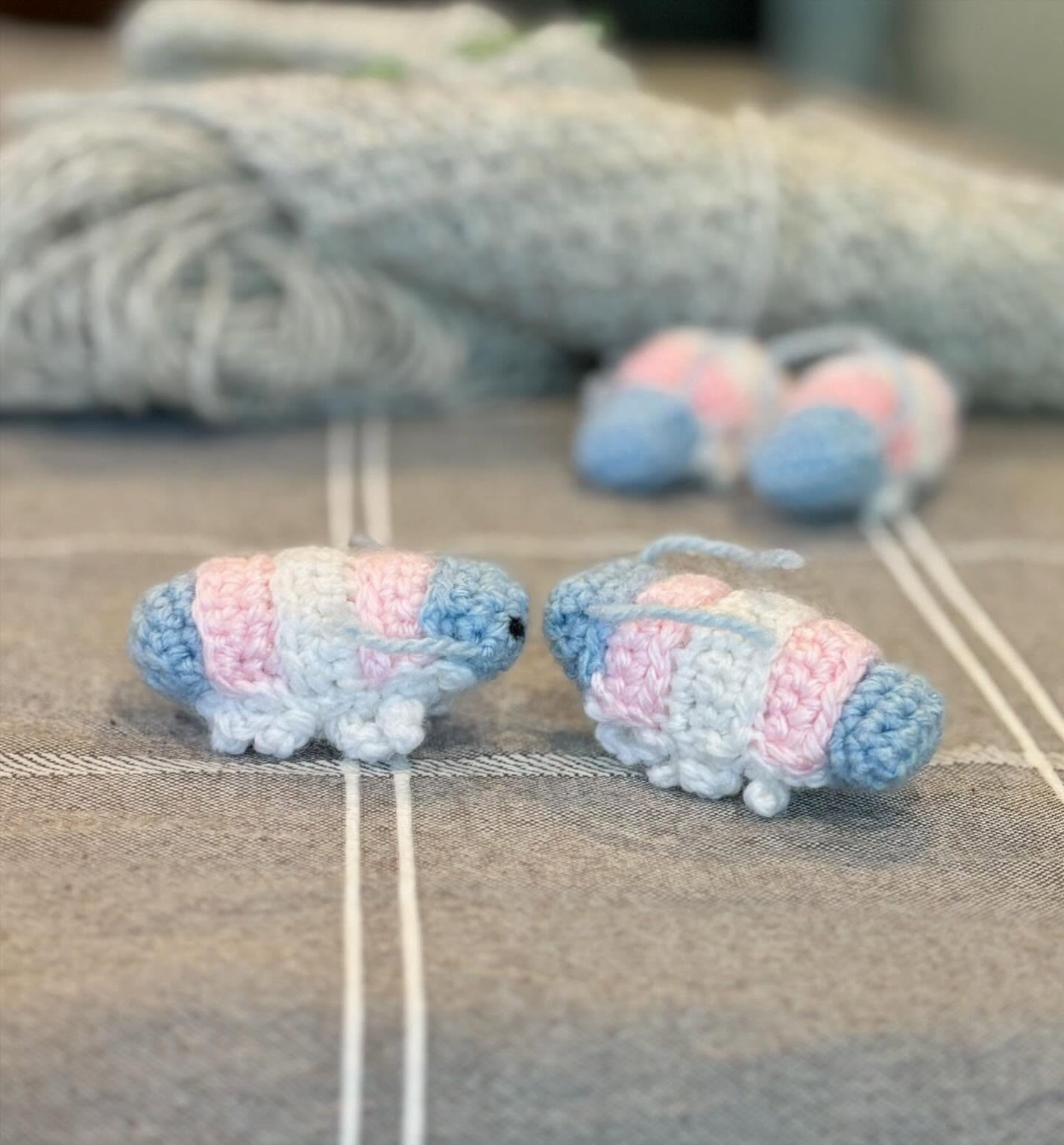 Foreground: two small crocheted isopod bugs face to face, each shell/exoskeleton section is a different colour of the trans flag. Background: grey yarn with two more isopods of the same design facing forward.