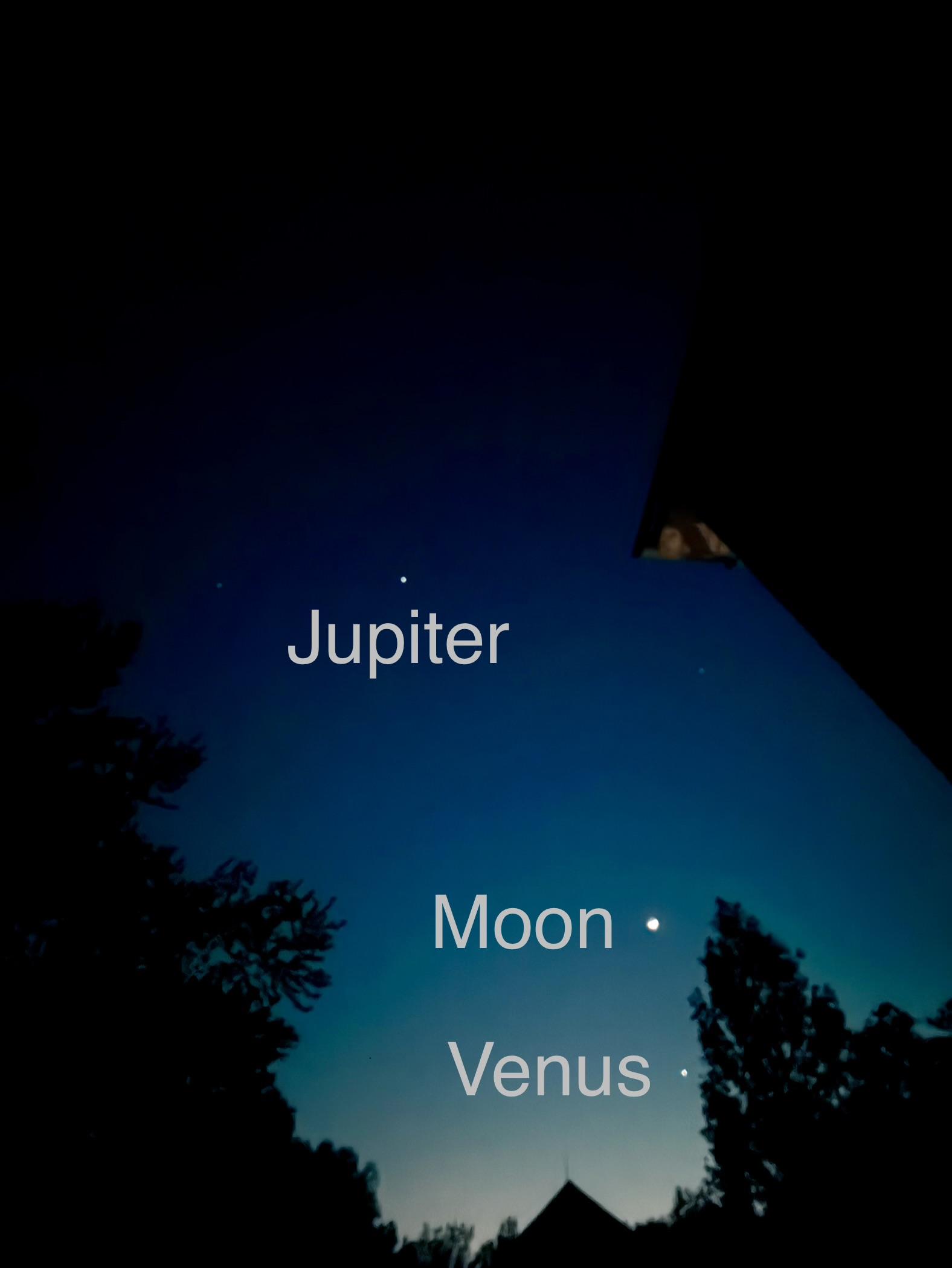 Photograph of the evening sky pointing west after dusk. The moon, Venus, and Jupiter are labeled in the dark blue sky, with the dark shape of trees and a barn framing the bottom of the image.