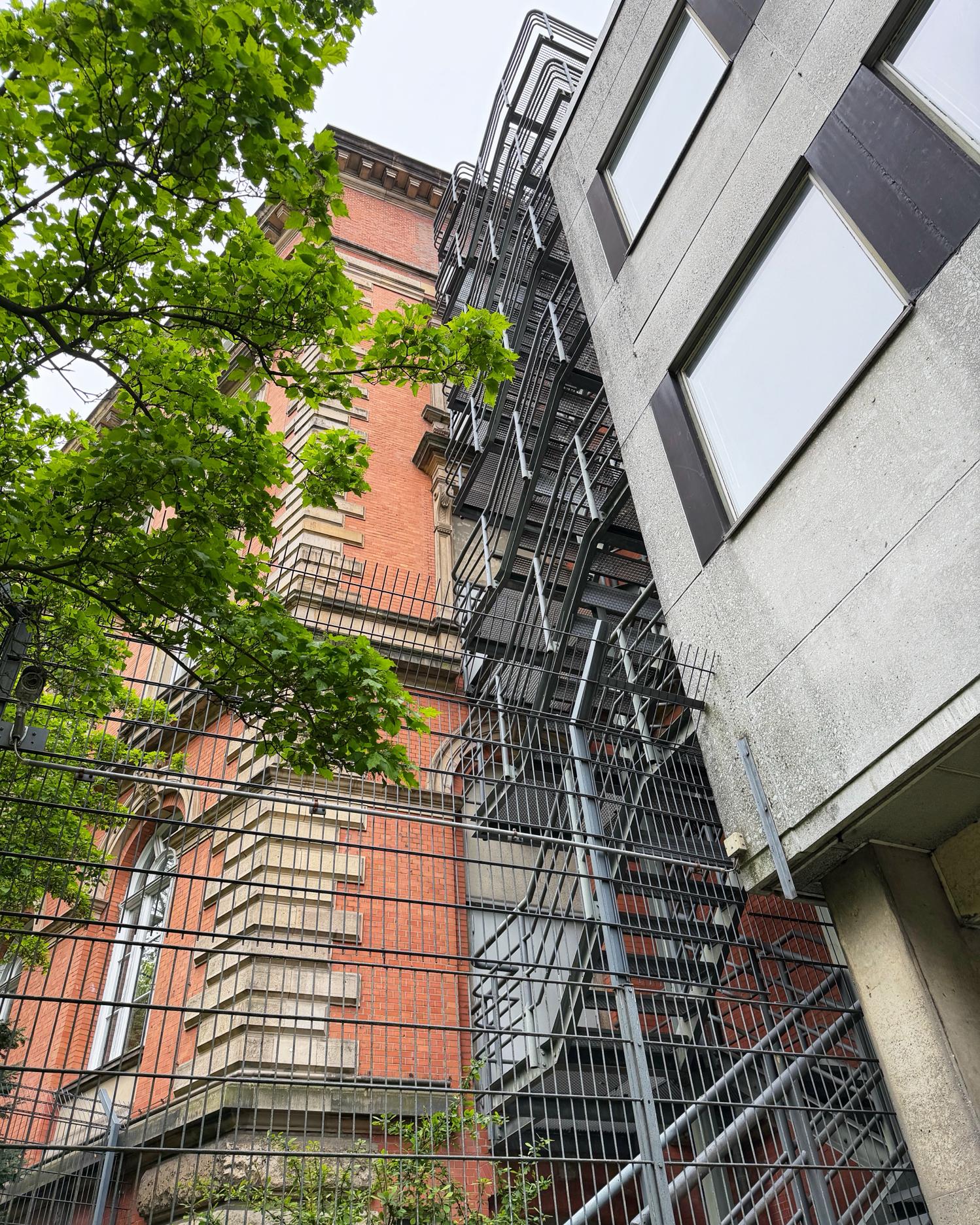 Metalltreppen und Gitterstrukturen verlaufen an der Fassade eines mehrstöckigen Gebäudes mit roten Ziegeln und hellen Steinelementen. Im Vordergrund steht ein grüner Laubbaum, rechts grenzt ein modernes Gebäude mit grauer Fassade und dunklen Fensterrahmen an. Die Perspektive ist von unten nach oben gerichtet.