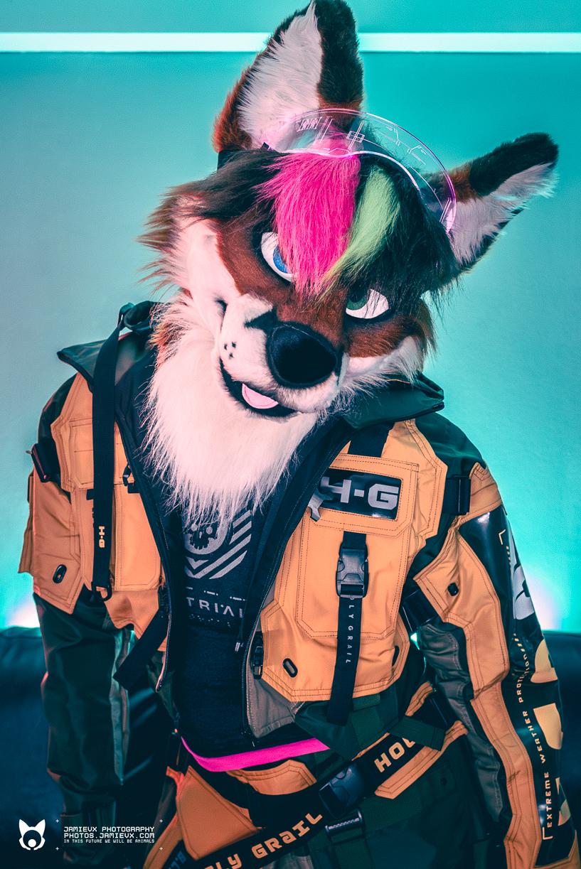 A photo of me wearing my fox fursuit head, and a yellow-orange and black techwear jacket. I am looking into the camera with a turquoise light shining behind me, contrasting with the yellows and oranges of my outfit.