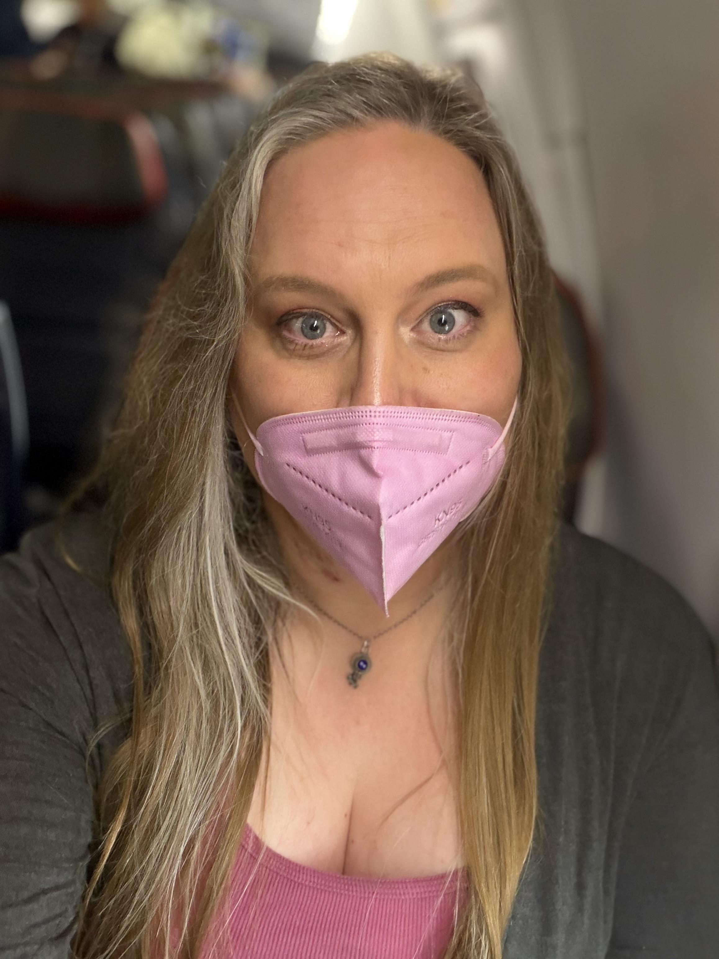 Selfie of a white trans woman on an airplane wearing a pink face mask.