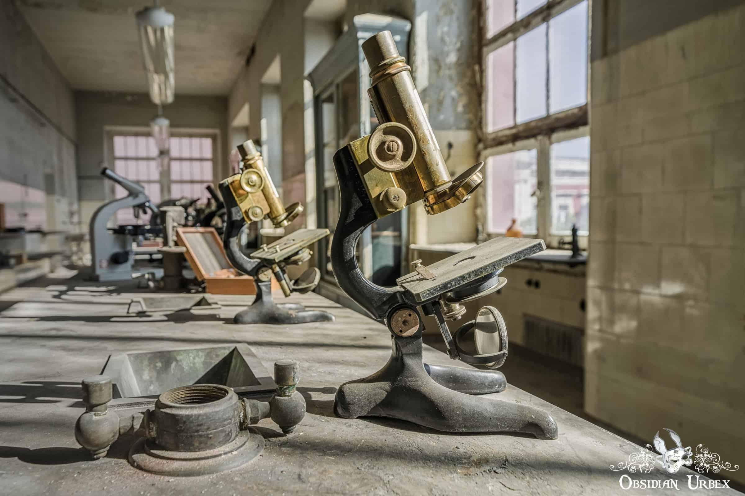 A vintage brass microscope sits on a dusty lab counter in an abandoned medical research institute, with other equipment