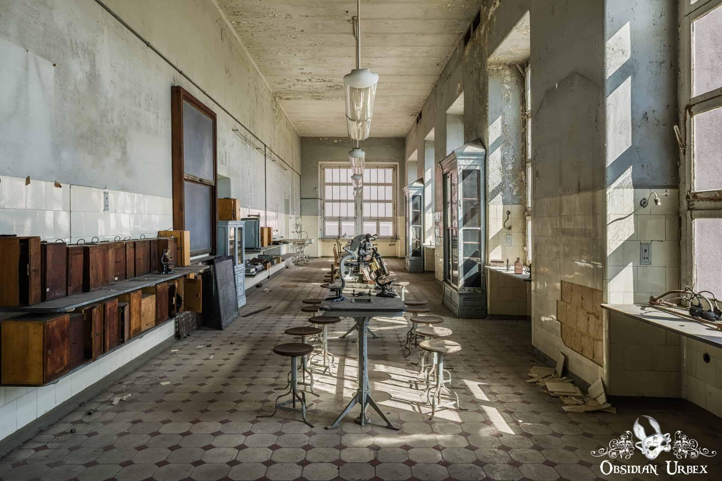 An abandoned medical teaching laboratory with peeling paint, long tables, stools, and vintage microscopes.